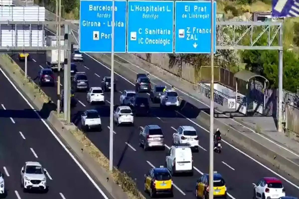 Tráfico moderado en una autopista de varios carriles con señales azules que indican direcciones hacia Barcelona, L'Hospitalet y El Prat, además de la presencia de taxis y vehículos particulares.