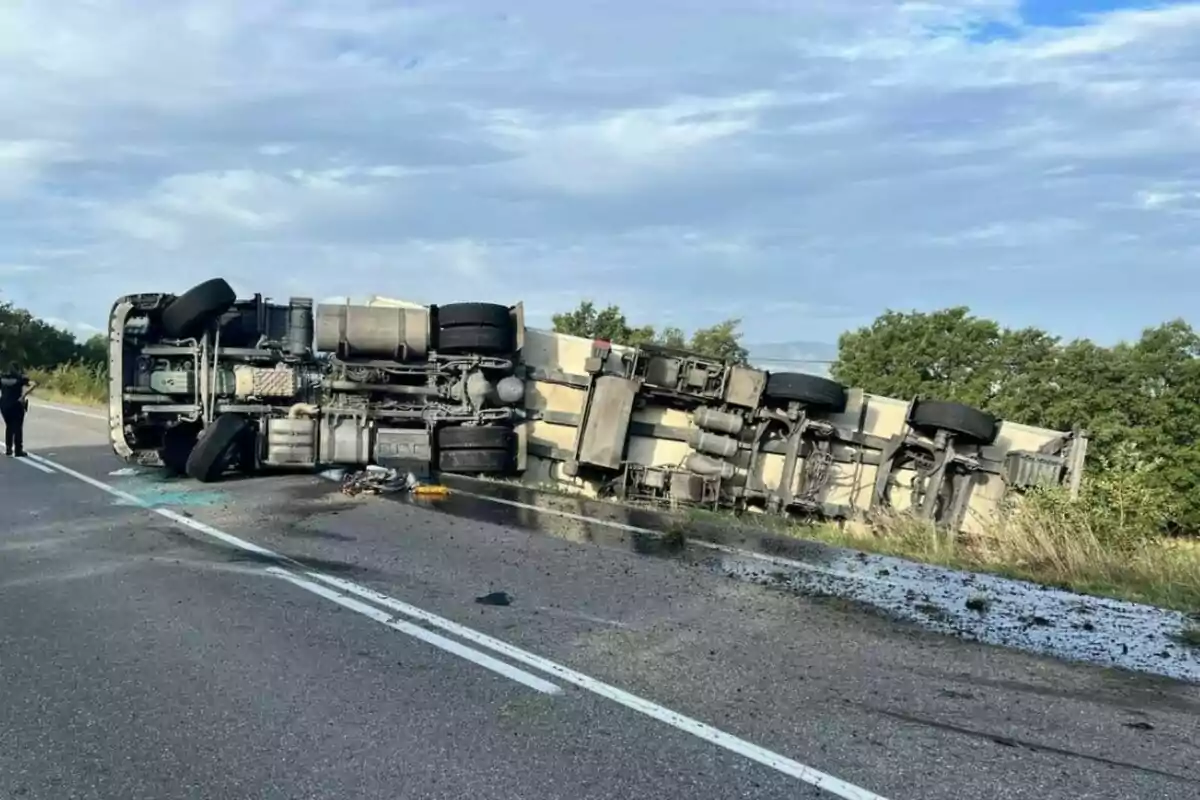 Camió de càrrega bolcat de costat en una carretera amb runa i líquids vessats a l’asfalt mentre una persona observa l’escena.