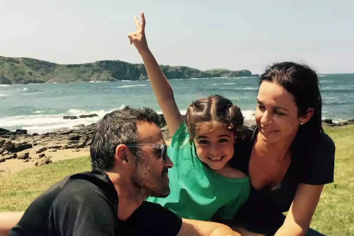 Familia sonriente sentada en el césped junto al mar con una niña levantando la mano haciendo el signo de la paz Familia sonriente sentada en el césped junto al mar con una niña levantando la mano haciendo el signo de la paz