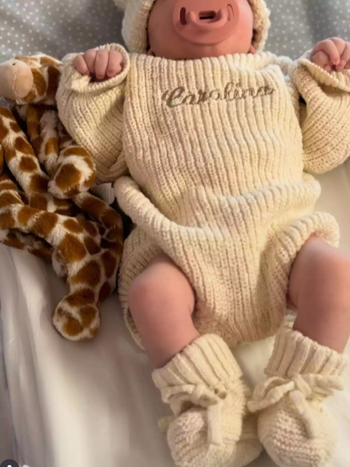 Bebé acostado usando un conjunto de punto color crema con el nombre Carolina bordado en el pecho, acompañado de un chupete rosa y un peluche de jirafa a su lado. Bebé acostado usando un conjunto de punto color crema con el nombre Carolina bordado en el pecho, acompañado de un chupete rosa y un peluche de jirafa a su lado.