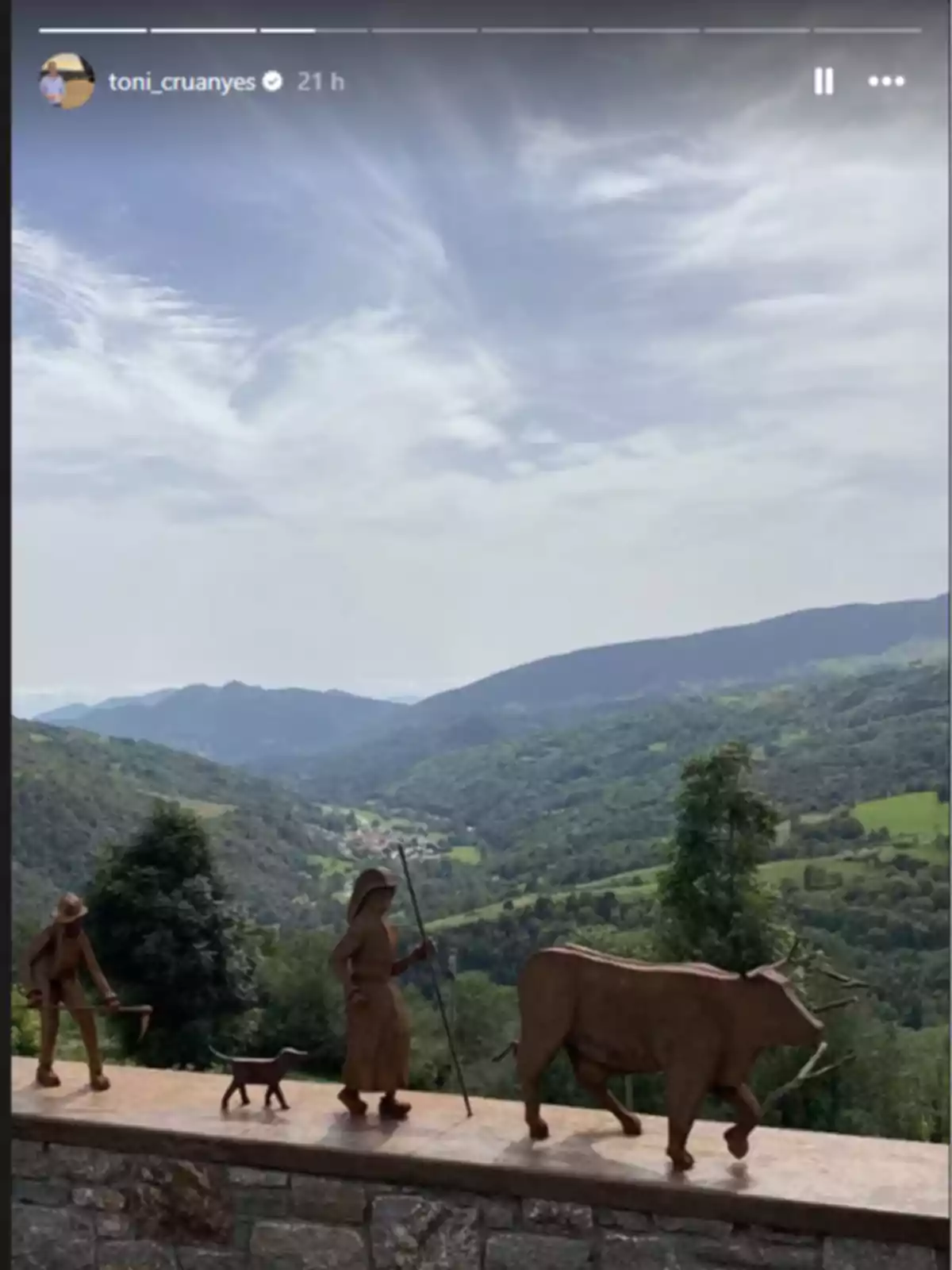 Escultura de figures rurals amb un bou i un gos sobre un mur de pedra amb un paisatge de muntanyes i valls verdes al fons sota un cel parcialment ennuvolat Escultura de figures rurals amb un bou i un gos sobre un mur de pedra amb un paisatge de muntanyes i valls verdes al fons sota un cel parcialment ennuvolat