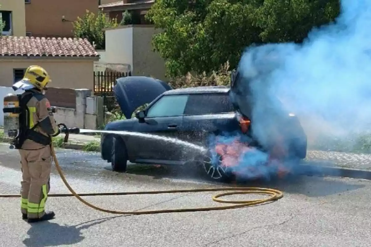 Un bomber apaga l’incendi en un cotxe negre estacionat al carrer mentre surt fum blau i flames de la part del darrere del vehicle.