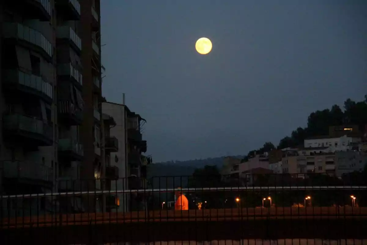 La luna llena brilla en el cielo nocturno sobre una ciudad con edificios y una persona caminando iluminada por una luz naranja