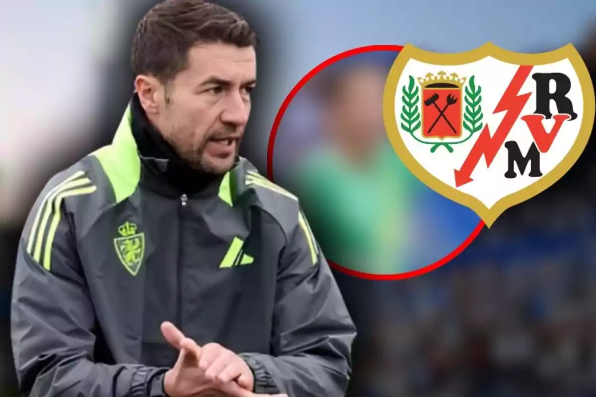 Entrenador de futbol amb jaqueta esportiva grisa i detalls verds al costat de l’escut del Rayo Vallecano sobre un fons desenfocat
