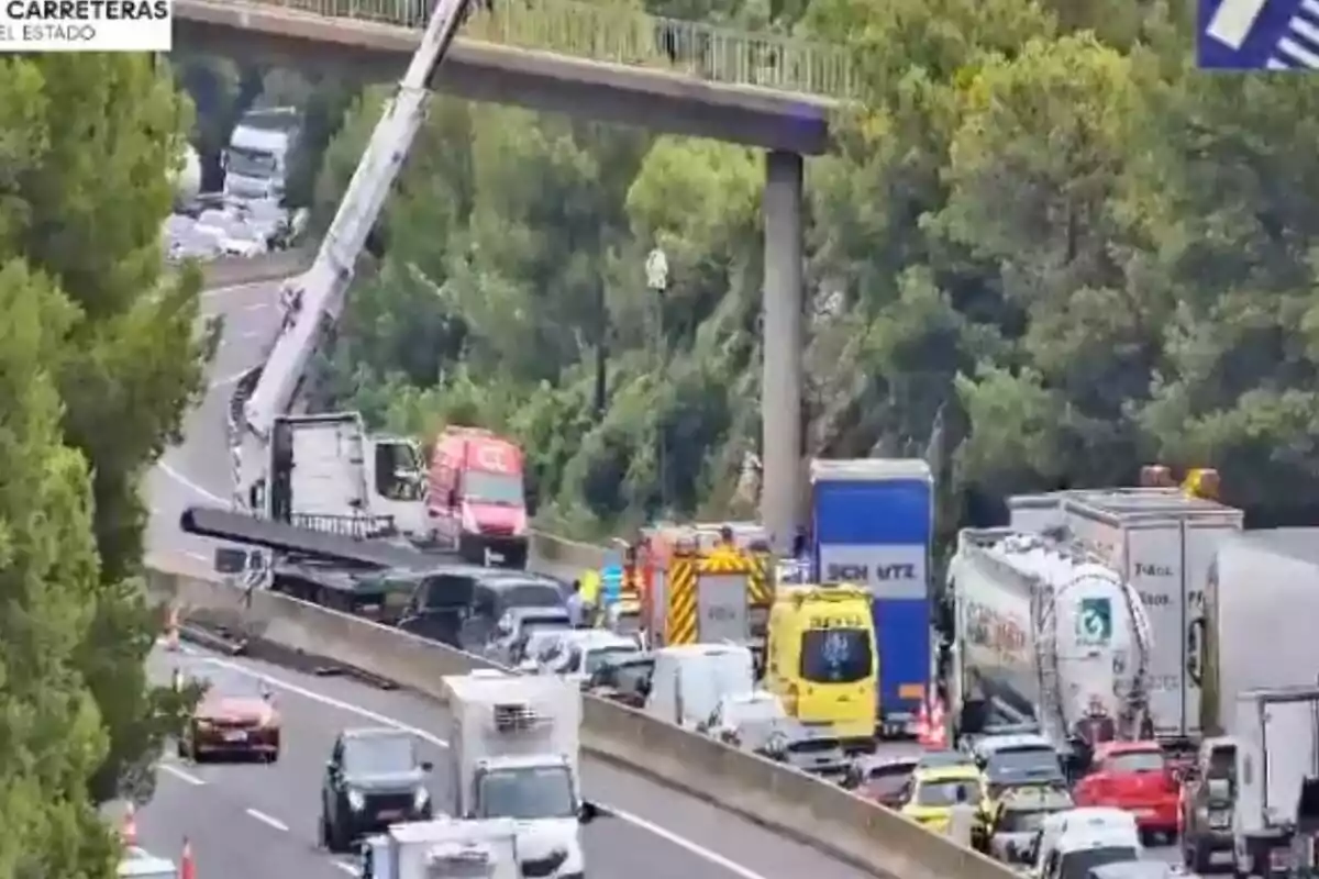 Accident en una autopista amb diversos vehicles d’emergència i trànsit aturat al costat d’un pont per a vianants envoltat d’arbres