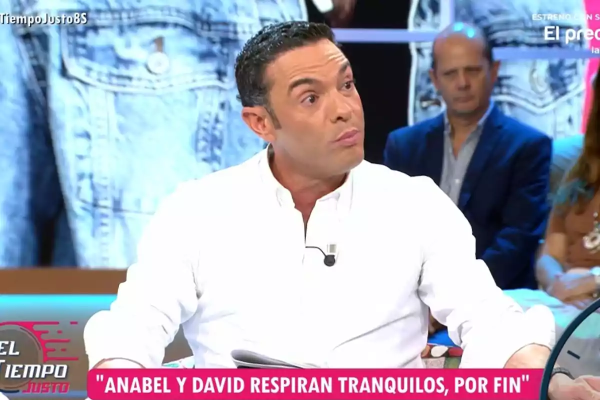 Home amb camisa blanca participant en un programa de televisió, amb expressió seriosa i un micròfon a la solapa, mentre el públic observa al fons i a la pantalla apareix un titular sobre l’Anabel i en David. Home amb camisa blanca participant en un programa de televisió, amb expressió seriosa i un micròfon a la solapa, mentre el públic observa al fons i a la pantalla apareix un titular sobre l’Anabel i en David.