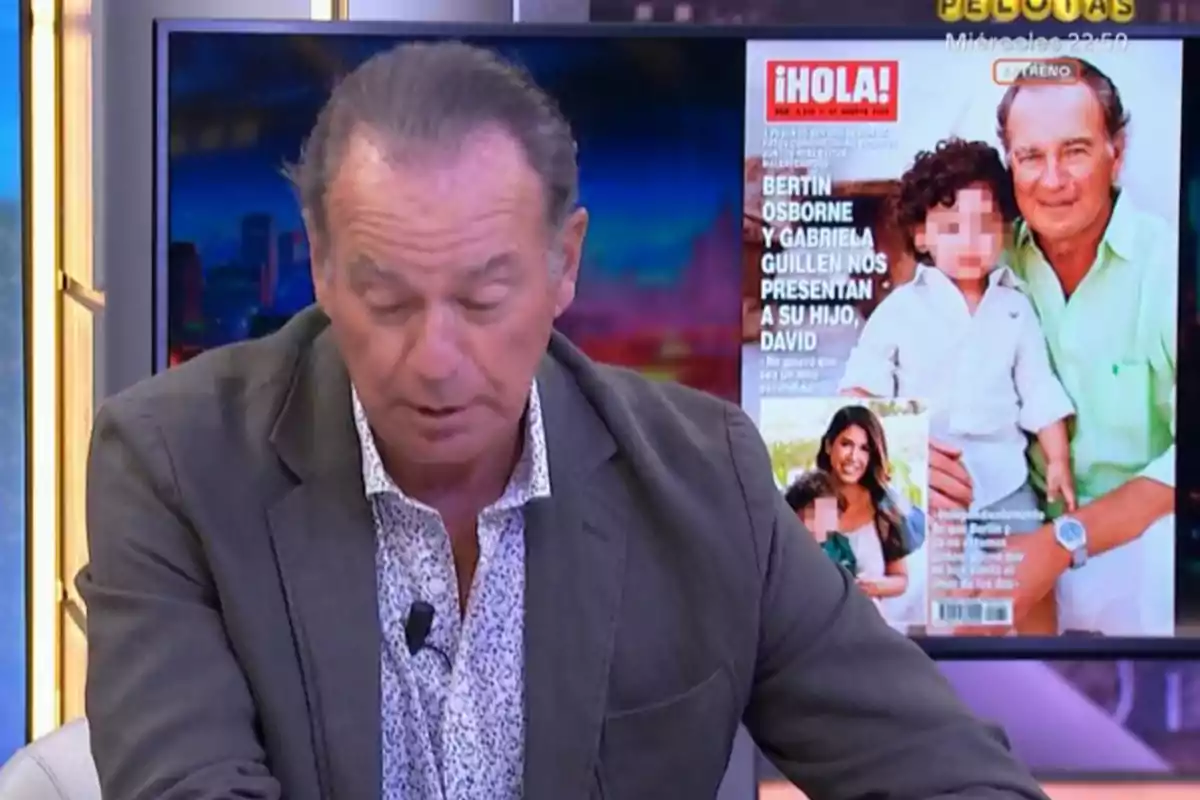Un home amb els cabells recollits i jaqueta grisa està assegut en un estudi de televisió; al fons es veu una pantalla amb la portada d’una revista on apareix el mateix home juntament amb un nen i una dona. Un home amb els cabells recollits i jaqueta grisa està assegut en un estudi de televisió; al fons es veu una pantalla amb la portada d’una revista on apareix el mateix home juntament amb un nen i una dona.