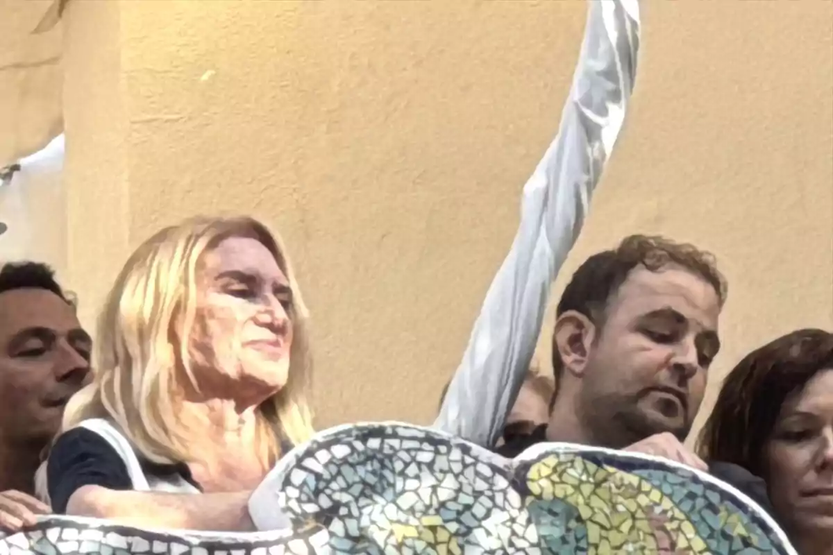 Varias personas están reunidas al aire libre junto a una estructura decorada con mosaicos, una mujer rubia está en primer plano y un hombre levanta el brazo. Varias personas están reunidas al aire libre junto a una estructura decorada con mosaicos, una mujer rubia está en primer plano y un hombre levanta el brazo.