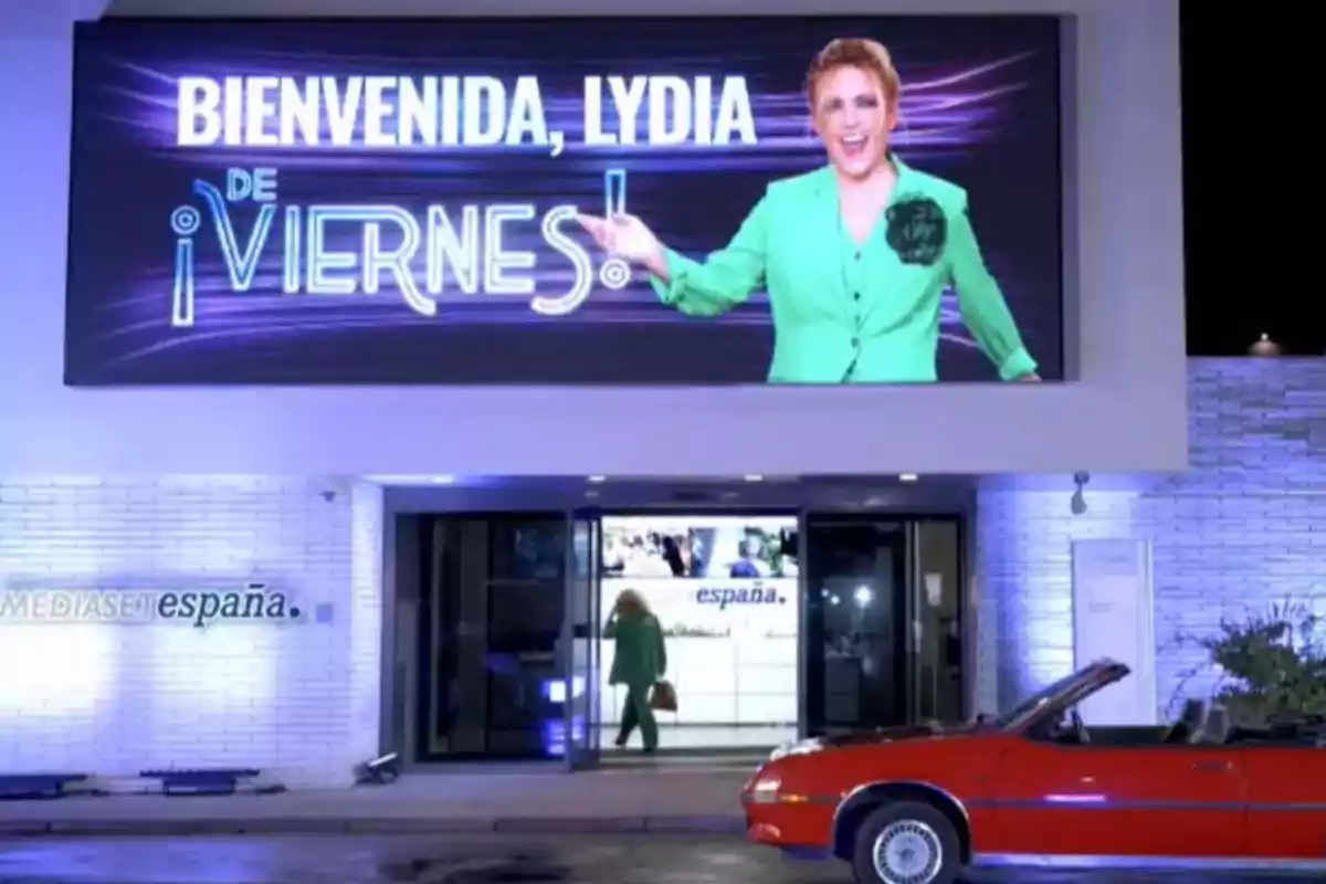 Una dona amb vestit verd apareix en una pantalla gegant sobre l’entrada d’un edifici amb el rètol “benvinguda Lydia de divendres” mentre un cotxe vermell descapotable està estacionat davant la porta. Una dona amb vestit verd apareix en una pantalla gegant sobre l’entrada d’un edifici amb el rètol “benvinguda Lydia de divendres” mentre un cotxe vermell descapotable està estacionat davant la porta.