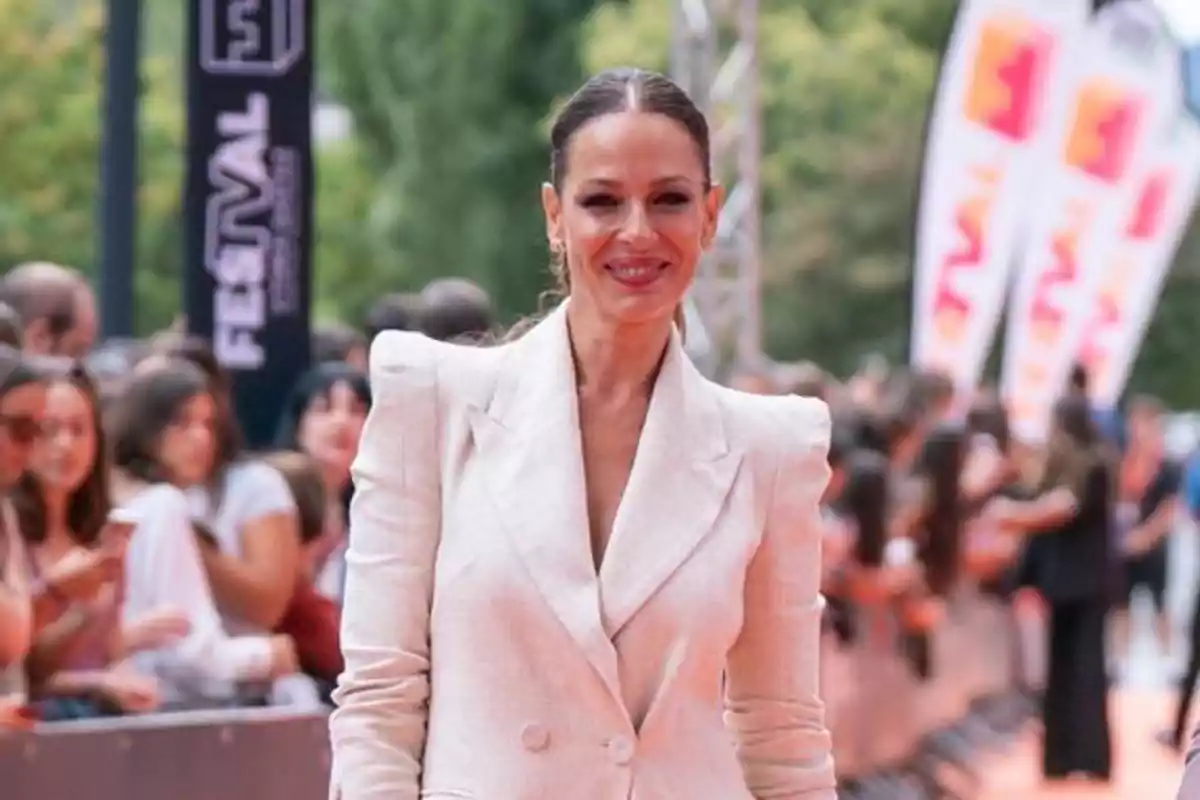 Eva González con saco blanco sonríe mientras camina por una alfombra naranja rodeada de personas y banderas en un evento al aire libre Eva González con saco blanco sonríe mientras camina por una alfombra naranja rodeada de personas y banderas en un evento al aire libre