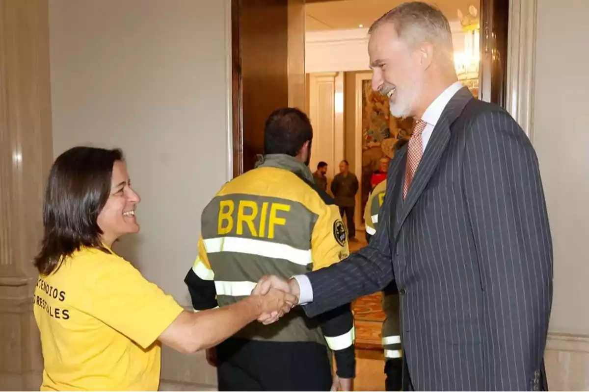 A man in a suit waves to a woman in a yellow T-jersey while other people in wildfire brigade uniforms are in the background. A man in a suit waves to a woman in a yellow T-jersey while other people in wildfire brigade uniforms are in the background.