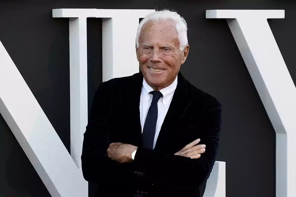 Giorgio Armani mayor de cabello blanco con traje oscuro y corbata posando sonriente frente a un fondo con letras grandes blancas Giorgio Armani mayor de cabello blanco con traje oscuro y corbata posando sonriente frente a un fondo con letras grandes blancas