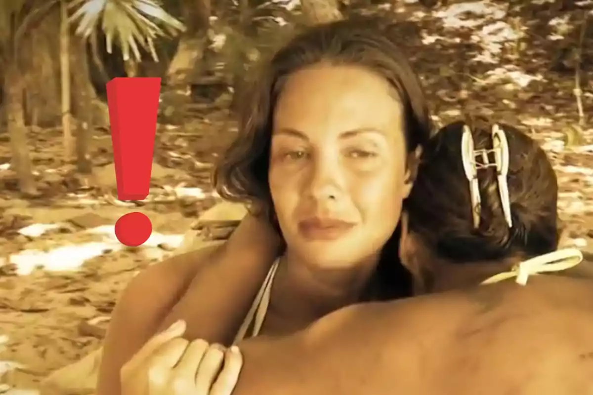 Dues dones s’abracen a la platja mentre una d’elles mira a la càmera amb expressió seriosa i un gran signe d’exclamació vermell apareix a l’esquerra.