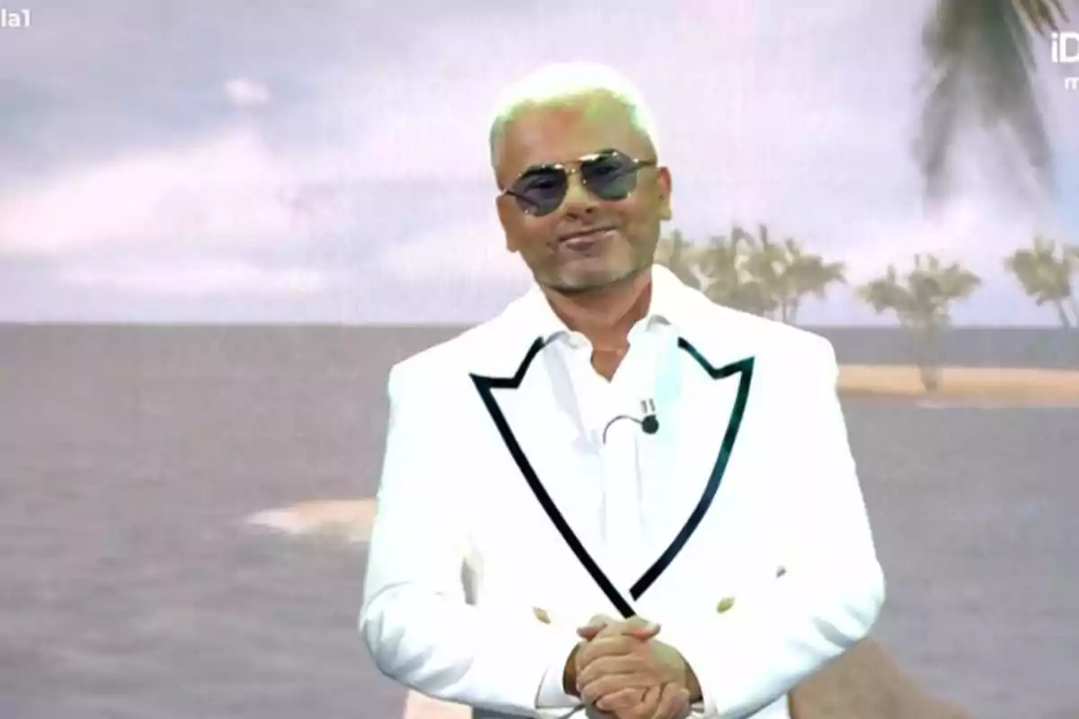 Jorge Javier Vázquez con gafas de sol y traje blanco posando frente a un fondo de playa con palmeras Jorge Javier Vázquez con gafas de sol y traje blanco posando frente a un fondo de playa con palmeras