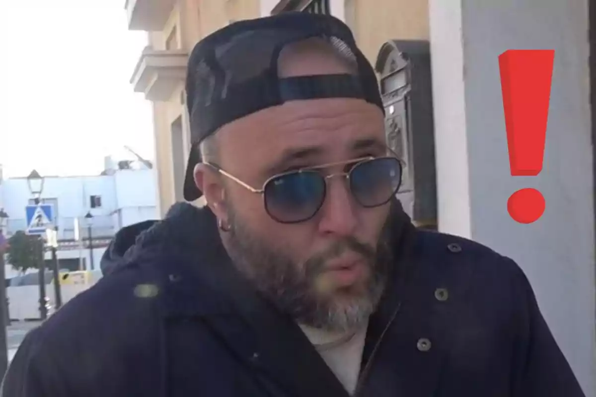 Kiko Rivera con barba y gafas de sol usando gorra hacia atrás, con un gesto de sorpresa y un signo de exclamación rojo al fondo