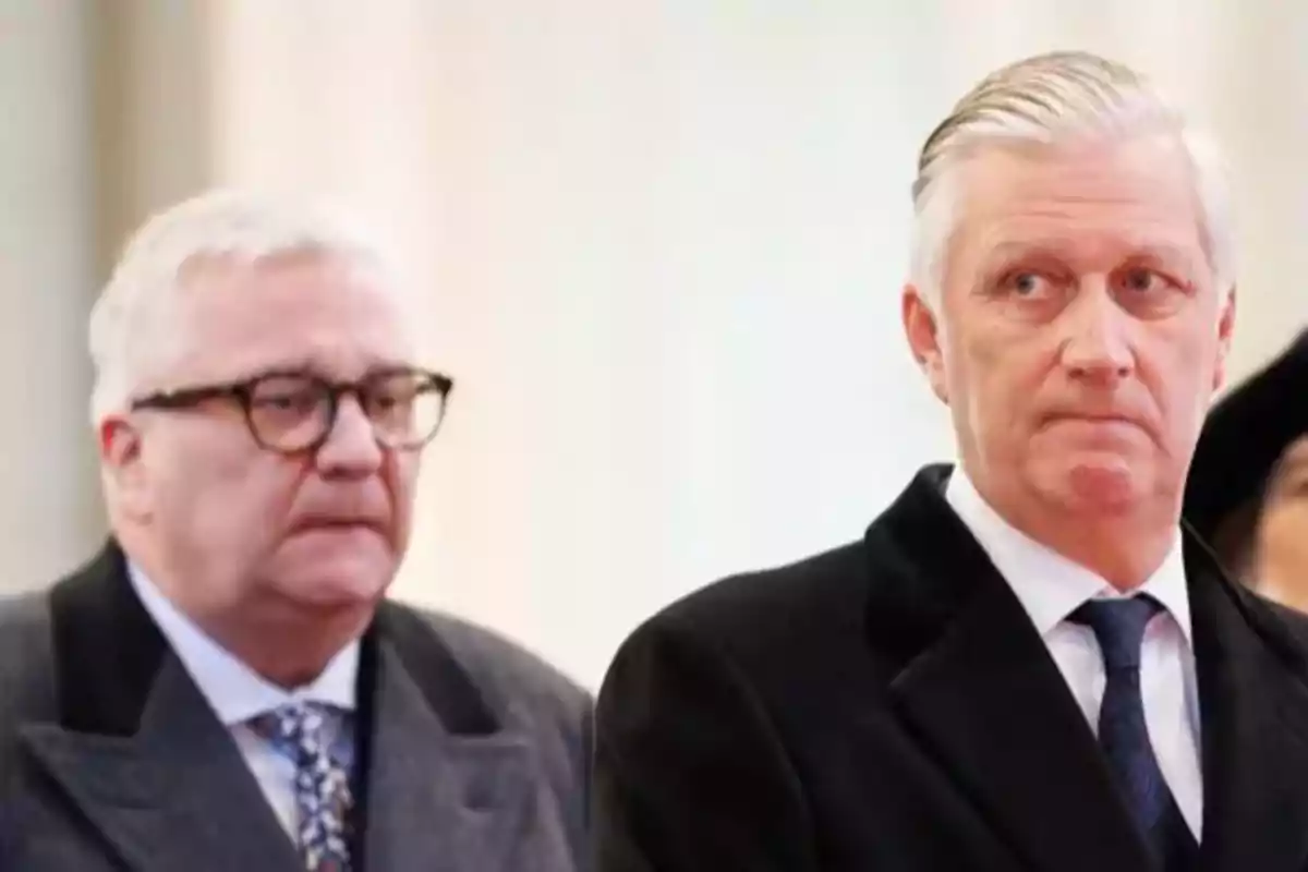 Lorenzo y Felipe de Bélgica mayores vestidos con traje formal miran hacia el frente con expresiones serias en un entorno interior iluminado. Lorenzo y Felipe de Bélgica mayores vestidos con traje formal miran hacia el frente con expresiones serias en un entorno interior iluminado.