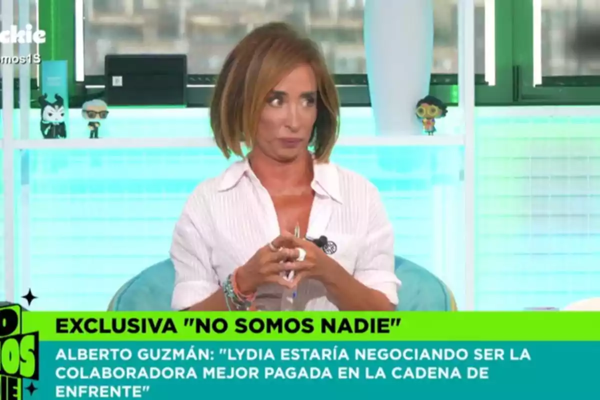 Dona amb cabell curt i ros asseguda en un plató de televisió amb expressió seriosa, porta una camisa blanca i darrere d’ella hi ha prestatgeries amb figures decoratives i una finestra; a la part inferior apareix un requadre verd amb text d’un programa de televisió. Dona amb cabell curt i ros asseguda en un plató de televisió amb expressió seriosa, porta una camisa blanca i darrere d’ella hi ha prestatgeries amb figures decoratives i una finestra; a la part inferior apareix un requadre verd amb text d’un programa de televisió.
