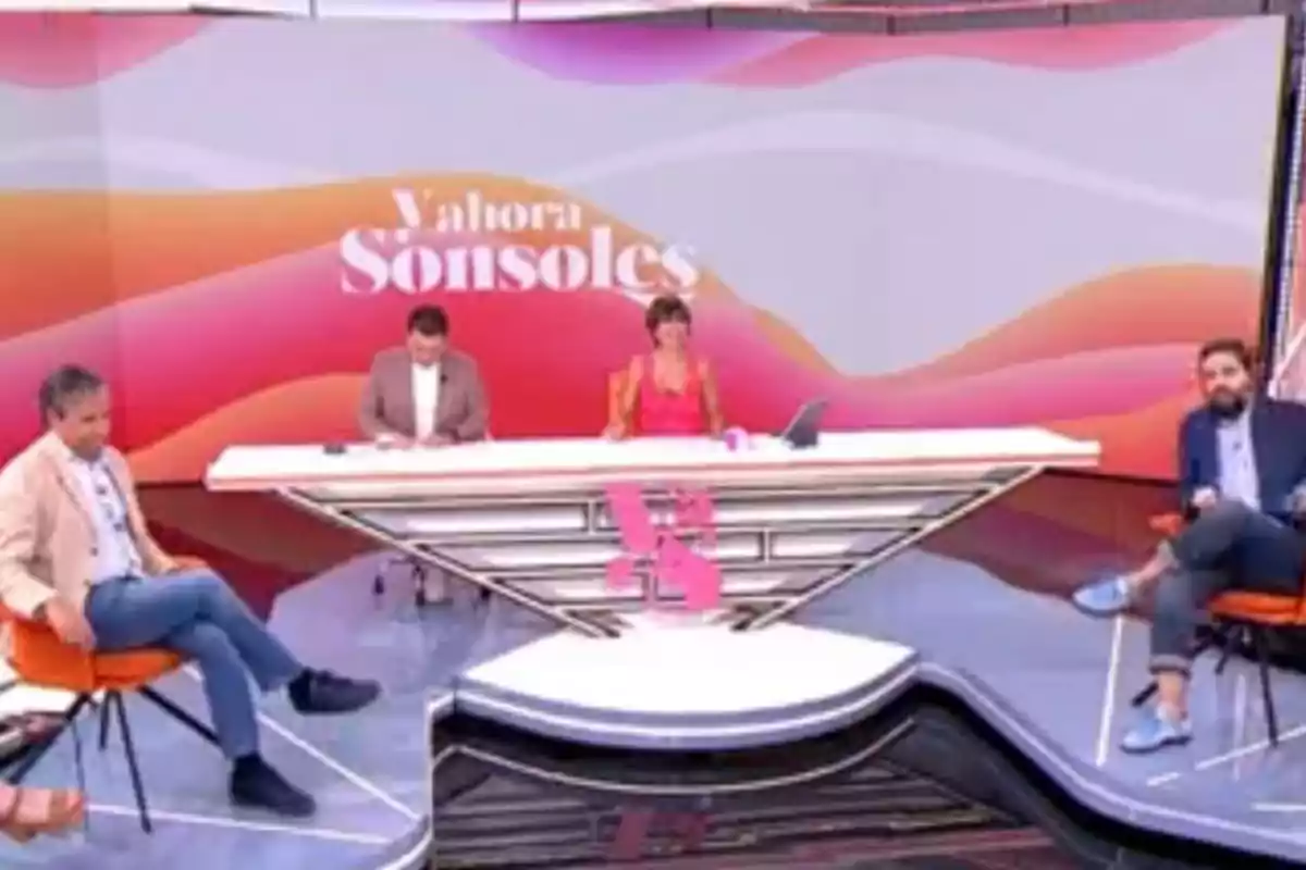 Un set de televisión con cuatro personas sentadas en un programa llamado Y ahora Sonsoles, con fondo de colores cálidos y una mesa blanca moderna. Un set de televisión con cuatro personas sentadas en un programa llamado Y ahora Sonsoles, con fondo de colores cálidos y una mesa blanca moderna.