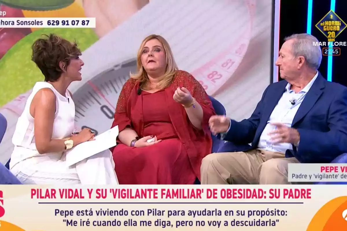Tres persones conversen en un programa de televisió, una dona amb vestit blanc, una dona amb vestit vermell i un home amb jaqueta blava, amb un fons d’una bàscula i una cinta mètrica, i un rètol a la part inferior que esmenta Pilar Vidal i el seu pare com a vigilant familiar de l’obesitat. Tres persones conversen en un programa de televisió, una dona amb vestit blanc, una dona amb vestit vermell i un home amb jaqueta blava, amb un fons d’una bàscula i una cinta mètrica, i un rètol a la part inferior que esmenta Pilar Vidal i el seu pare com a vigilant familiar de l’obesitat.