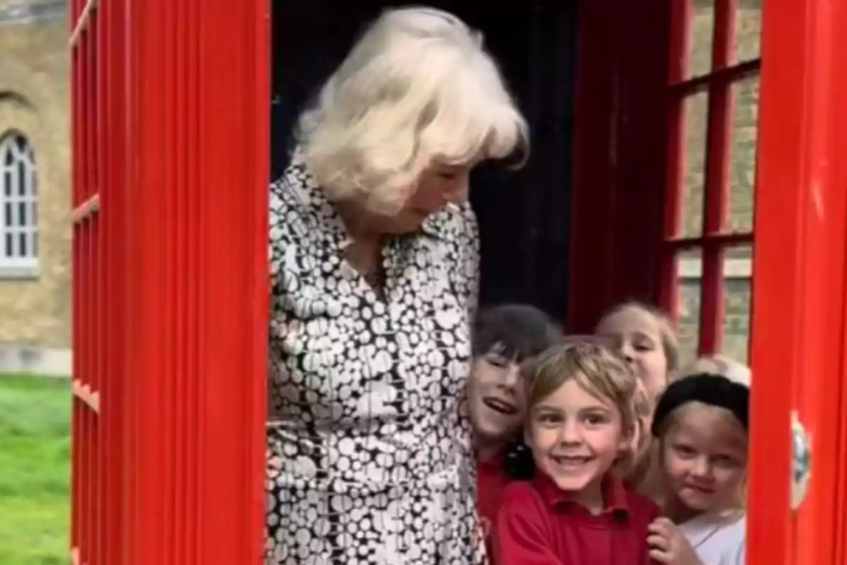 An elderly woman and several smiling children are inside a red phone booth. An elderly woman and several smiling children are inside a red phone booth.