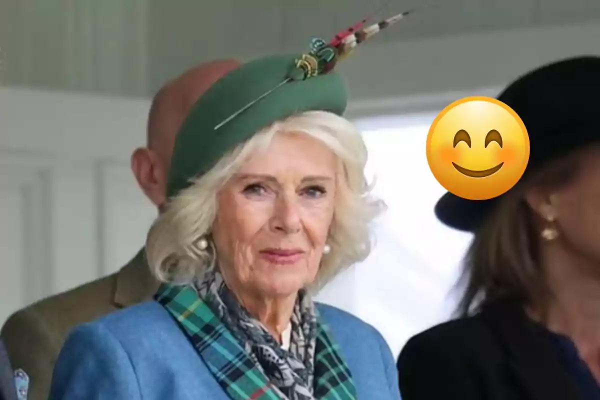 Short-haired blonde woman wearing a green hat decorated with feathers and a checkered scarf, accompanied by other people, one of whom has a smiling emoji covering their face.