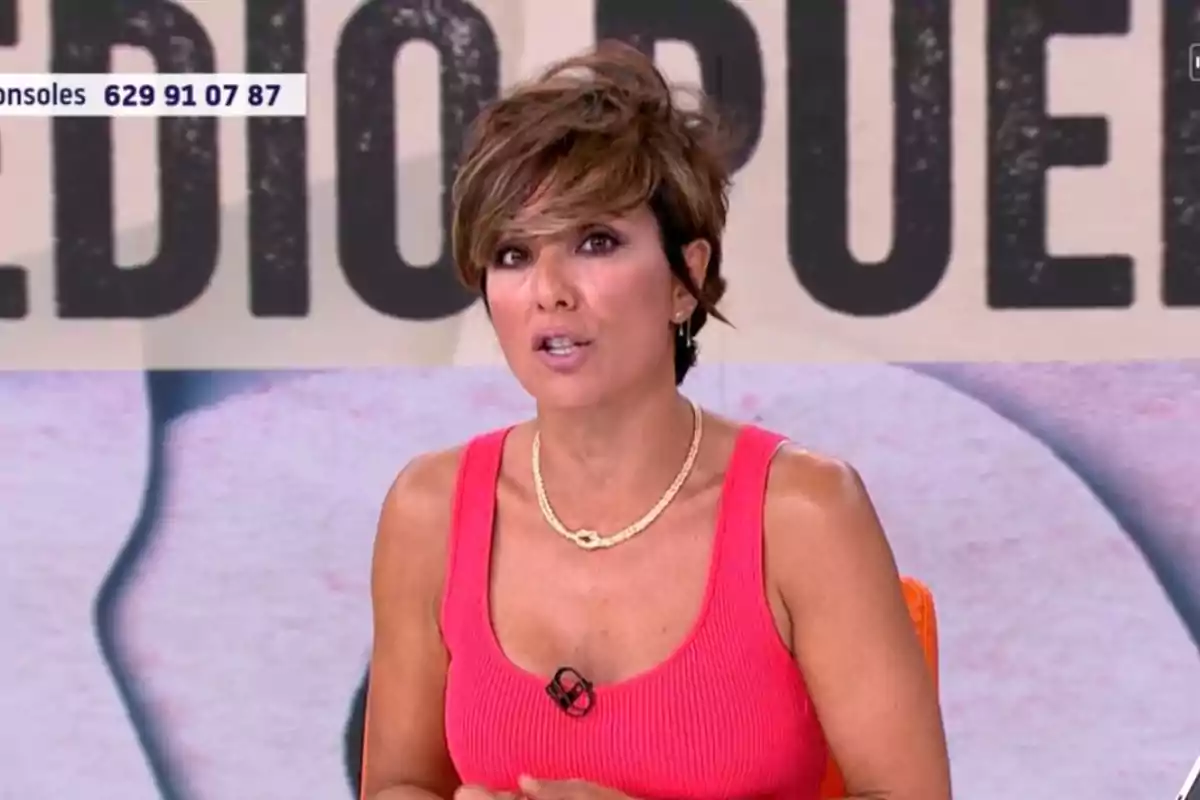 Sonsoles Ónega de cabello corto y castaño con vestido rosa y collar dorado hablando en un programa de televisión con fondo de letras grandes desenfocadas. Sonsoles Ónega de cabello corto y castaño con vestido rosa y collar dorado hablando en un programa de televisión con fondo de letras grandes desenfocadas.