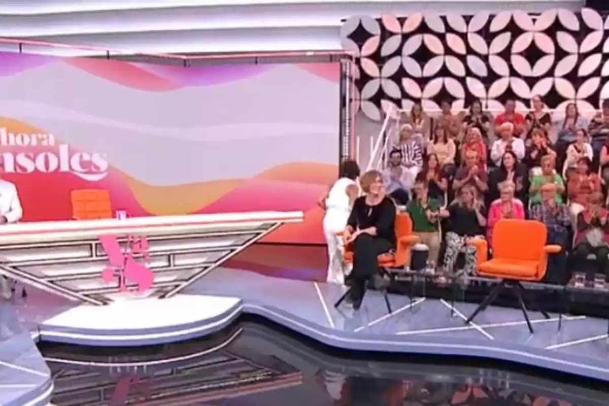 Plató de televisió amb públic assegut al fons, diverses persones en cadires taronges i una dona dreta vestida de blanc Plató de televisió amb públic assegut al fons, diverses persones en cadires taronges i una dona dreta vestida de blanc