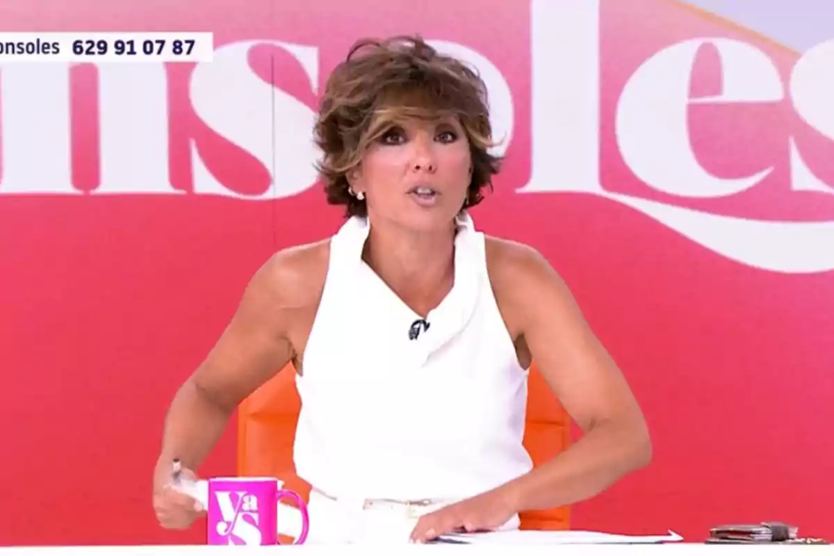 Presentadora de televisió asseguda en un plató amb fons vermell i blanc, vestida de blanc i amb una tassa rosa sobre la taula Presentadora de televisió asseguda en un plató amb fons vermell i blanc, vestida de blanc i amb una tassa rosa sobre la taula