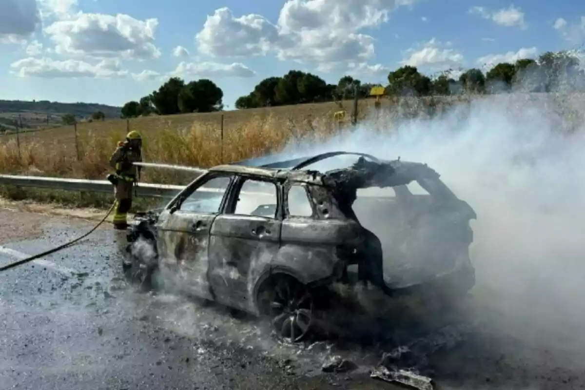 Un bomber apaga un cotxe completament calcinat en una carretera mentre surt fum del vehicle.