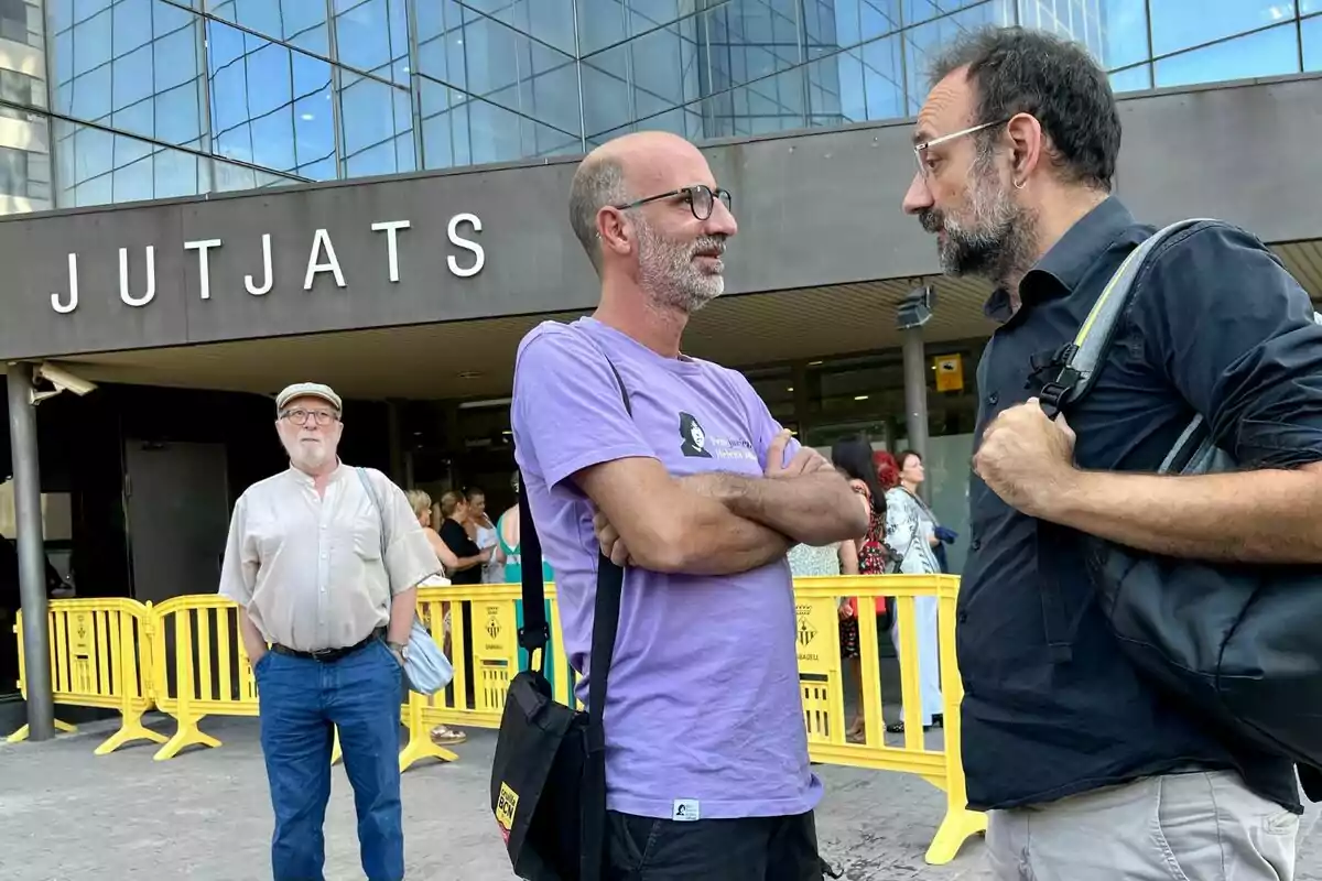 Tres hombres conversan frente a un edificio con el letrero JUTJATS y varias personas al fondo junto a vallas amarillas