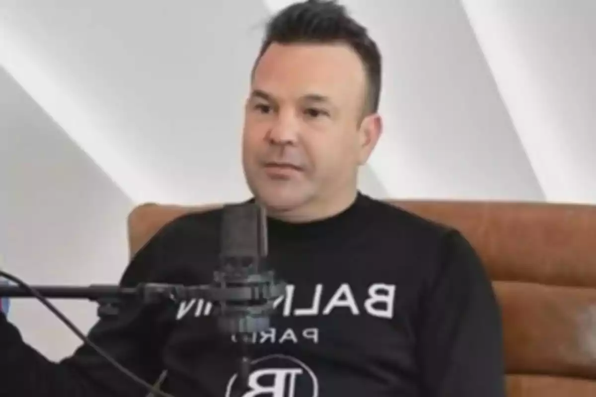 Hombre sentado en un sillón marrón usando un suéter negro con letras blancas frente a un micrófono durante una entrevista Hombre sentado en un sillón marrón usando un suéter negro con letras blancas frente a un micrófono durante una entrevista