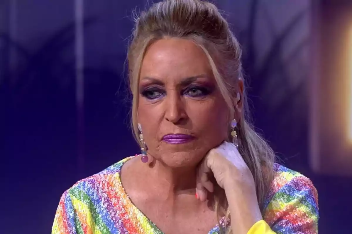 Dona rossa amb maquillatge cridaner i expressió seriosa, recolzant la barbeta a la mà i vestint una brusa de lluentons multicolor Dona rossa amb maquillatge cridaner i expressió seriosa, recolzant la barbeta a la mà i vestint una brusa de lluentons multicolor
