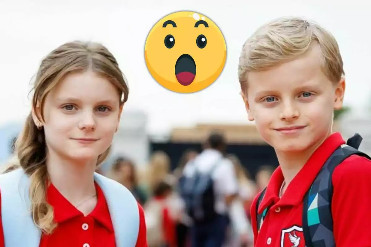 Gabriella i Jacques de Mònaco amb uniforme vermell i motxilles posen junts mentre al fons es veu un emoji de cara sorpresa