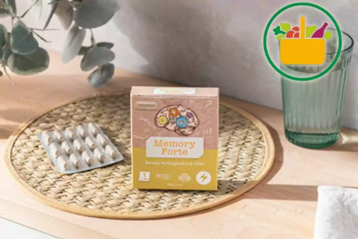 Caja de suplemento Memory Forte junto a un blíster de cápsulas sobre una bandeja tejida con un vaso de agua y plantas decorativas al fondo Caja de suplemento Memory Forte junto a un blíster de cápsulas sobre una bandeja tejida con un vaso de agua y plantas decorativas al fondo