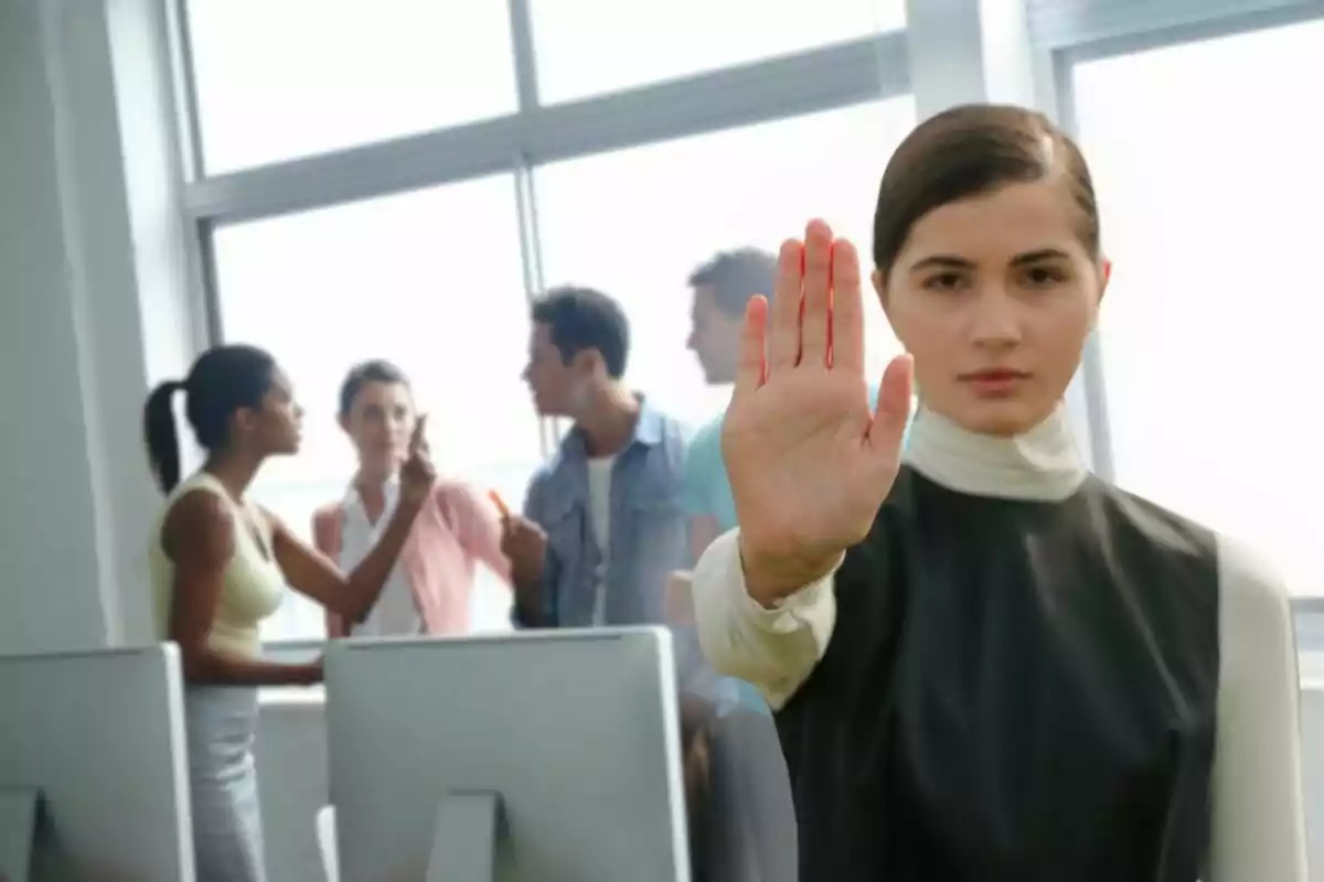 Mujer en primer plano levantando la mano en señal de alto mientras al fondo varias personas discuten en una oficina. Mujer en primer plano levantando la mano en señal de alto mientras al fondo varias personas discuten en una oficina.