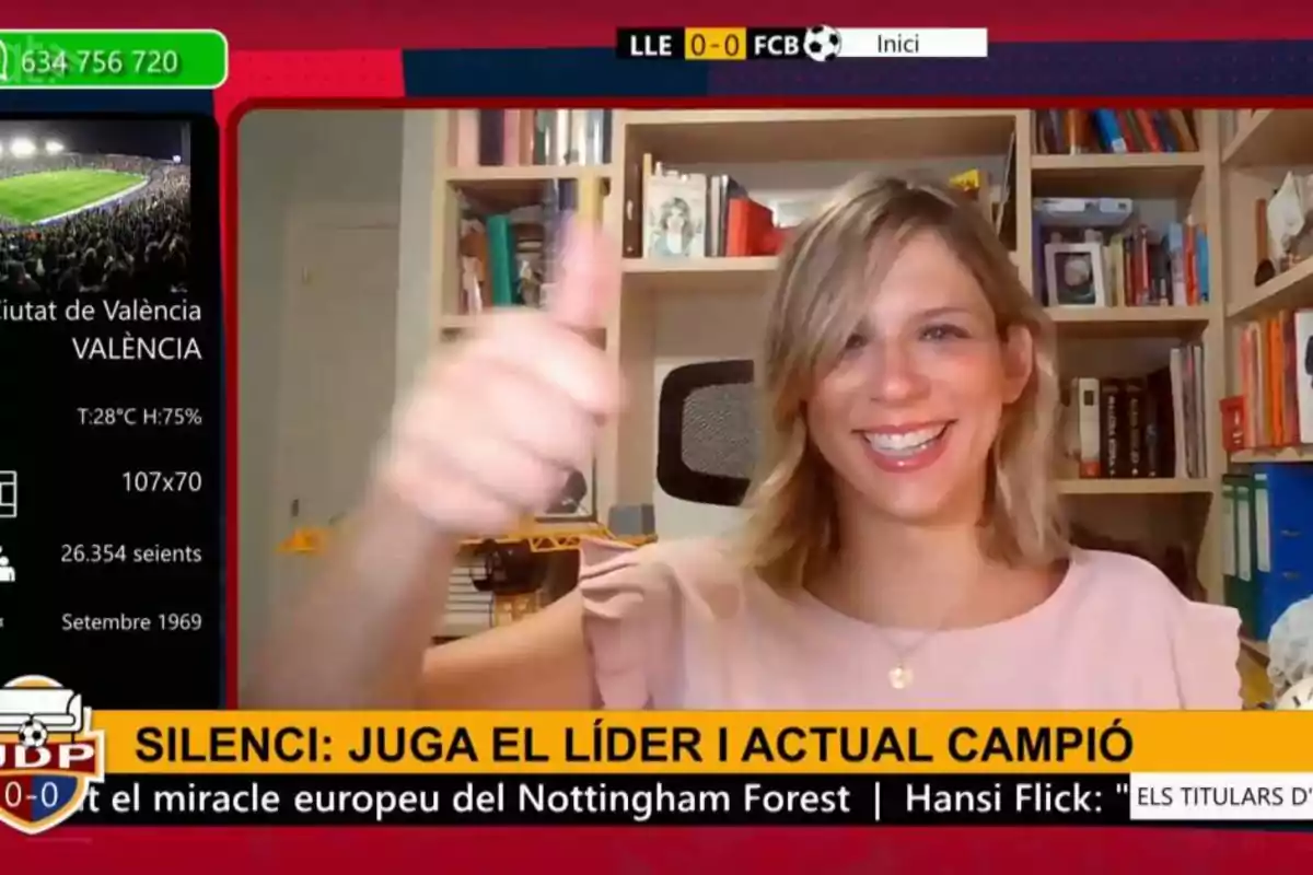 Mujer sonriente con el pulgar levantado durante una transmisión deportiva en la que se muestra información de un estadio y un marcador de fútbol en la pantalla. Mujer sonriente con el pulgar levantado durante una transmisión deportiva en la que se muestra información de un estadio y un marcador de fútbol en la pantalla.