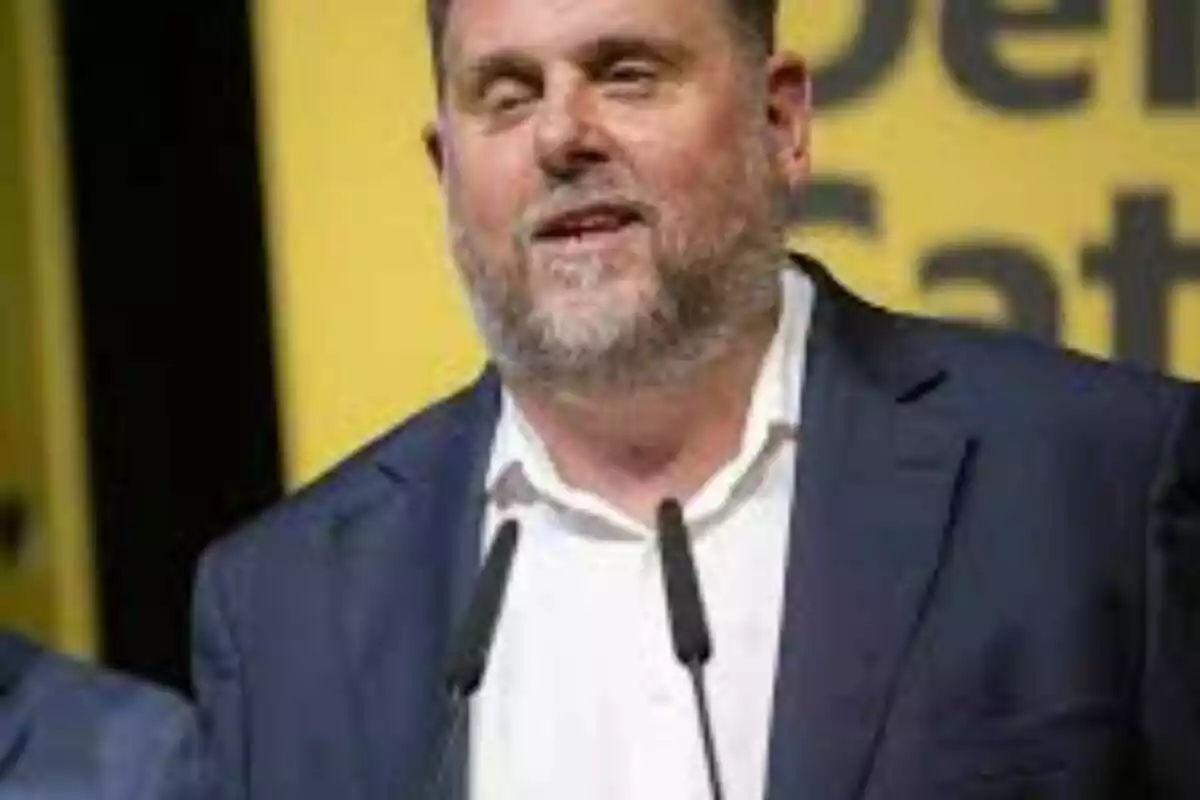 Hombre de barba y cabello corto con saco azul hablando frente a dos micrófonos y un fondo amarillo con letras negras Hombre de barba y cabello corto con saco azul hablando frente a dos micrófonos y un fondo amarillo con letras negras