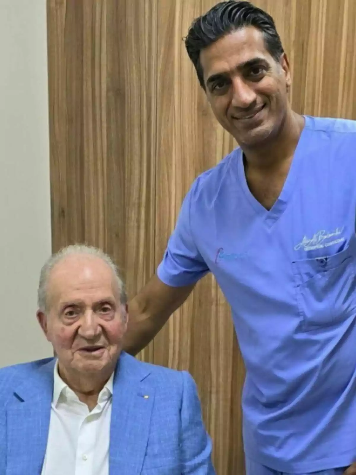 Juan Carlos I and Ali Albelooshi pose together in front of a wooden wall; one of them is sitting and wearing a light blue jacket while the other is standing, dressed in blue medical scrubs and smiling. Juan Carlos I and Ali Albelooshi pose together in front of a wooden wall; one of them is sitting and wearing a light blue jacket while the other is standing, dressed in blue medical scrubs and smiling.
