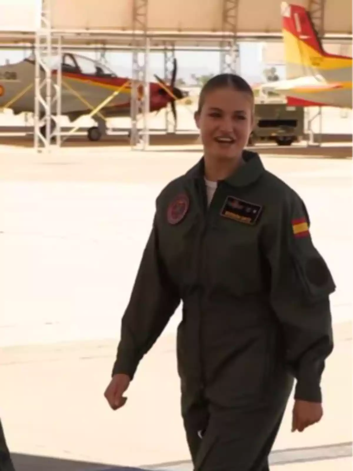 Princess Leonor in a green military flight suit walking in a hangar with airplanes in the background Princess Leonor in a green military flight suit walking in a hangar with airplanes in the background