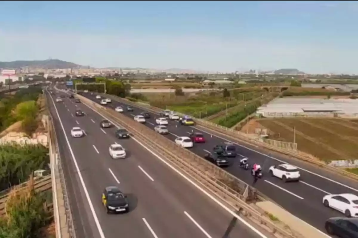 Vista aèria d’una autopista amb diversos cotxes i motos circulant en ambdós sentits, envoltada de camps i amb una ciutat al fons.