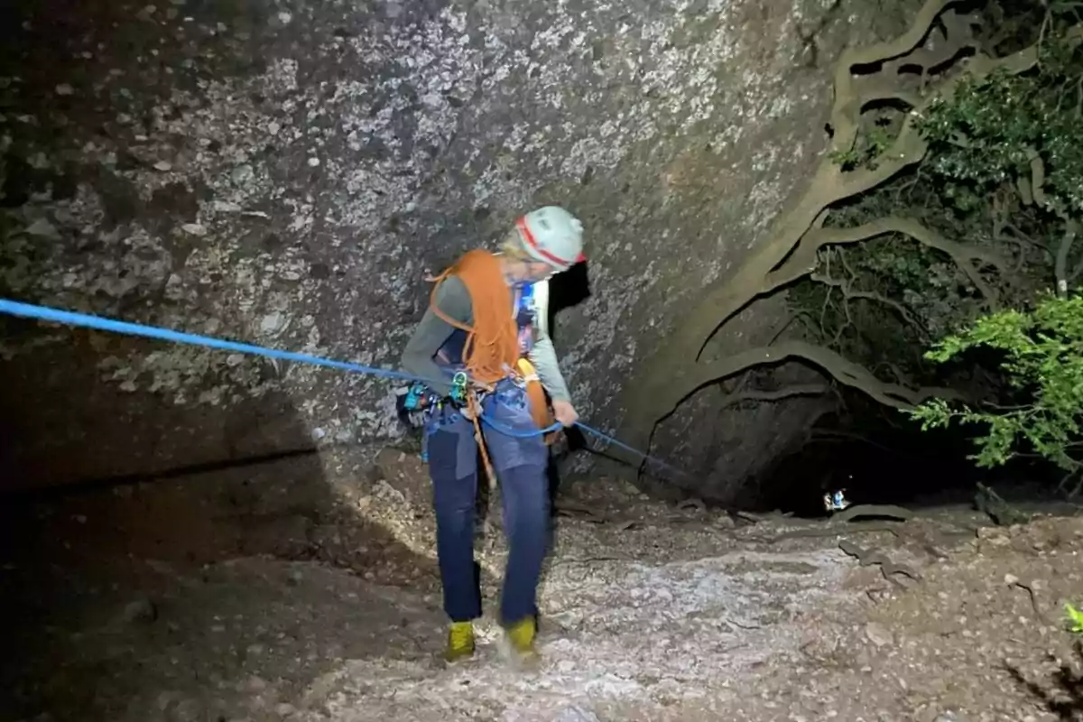 Persona amb casc i equip d’escalada descendint per una corda en una cova il·luminada artificialment, envoltada de parets rocoses i vegetació.