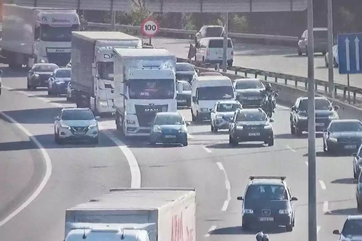 Tráfico denso en una autopista con varios coches, camiones y una motocicleta circulando en diferentes carriles bajo una señal de límite de velocidad de 100