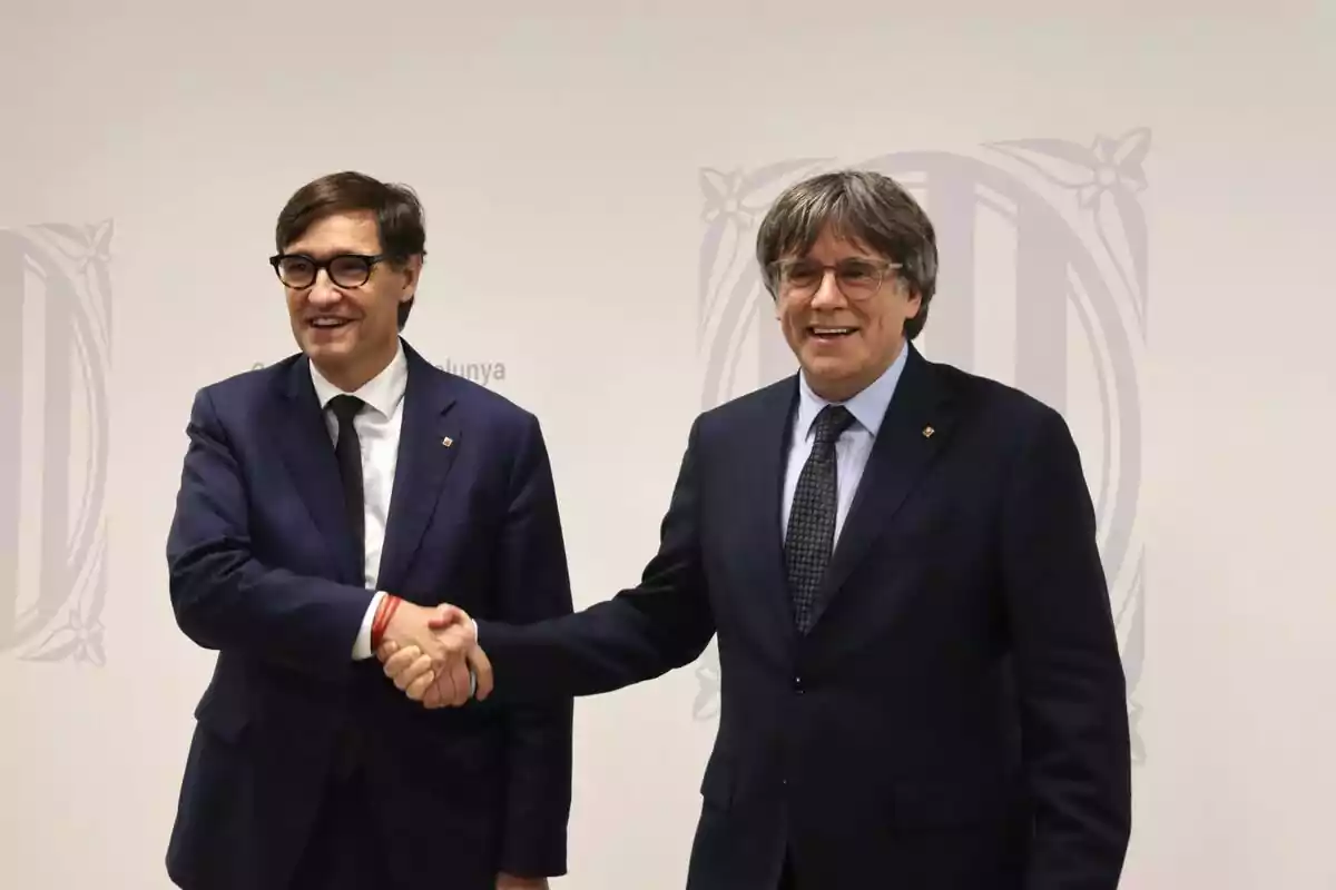 Dos homes amb ulleres i vestit fosc es donen la mà i somriuen davant d’un fons clar amb un logotip institucional.