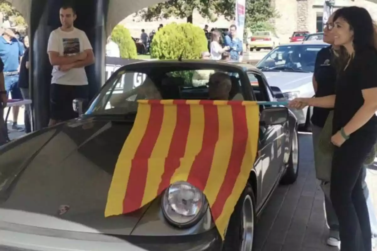 Una dona sosté una bandera groga amb franges vermelles davant d’un cotxe clàssic gris mentre diverses persones observen en un esdeveniment a l’aire lliure. Una dona sosté una bandera groga amb franges vermelles davant d’un cotxe clàssic gris mentre diverses persones observen en un esdeveniment a l’aire lliure.