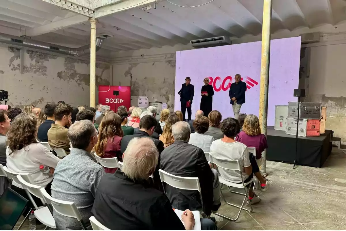 Un grup de persones assegudes en cadires blanques assisteix a una presentació en un espai industrial amb parets desgastades, mentre tres ponents estan drets en un escenari davant d’una pantalla gran amb el logotip de BCCIT.