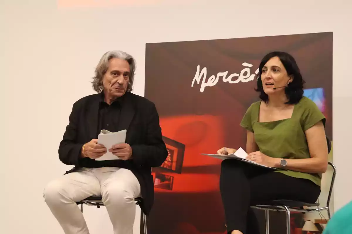 Dues persones assegudes en cadires, una sostenint papers i l'altra amb un micròfon, amb un cartell de fons que diu “Mercè”. Dues persones assegudes en cadires, una sostenint papers i l'altra amb un micròfon, amb un cartell de fons que diu “Mercè”.