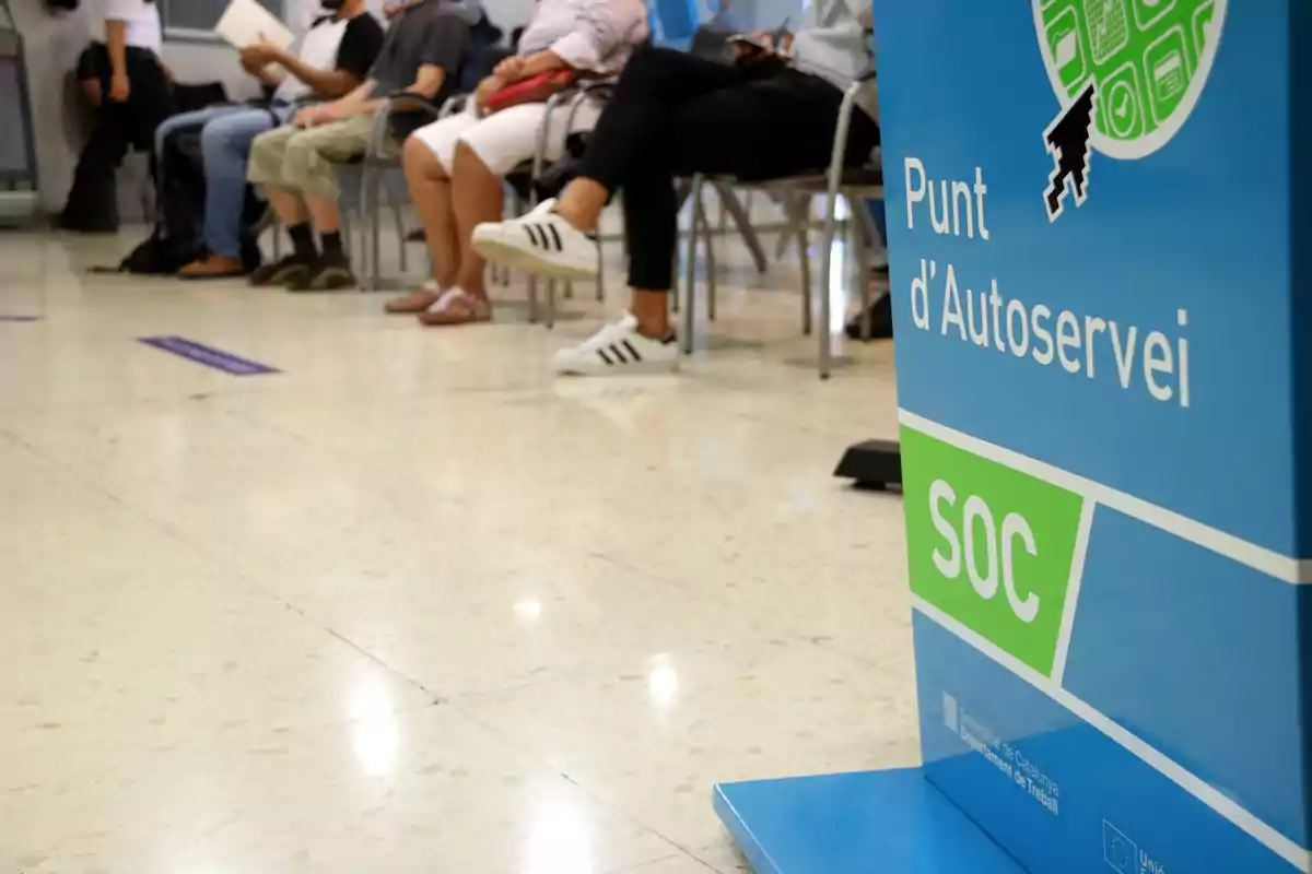 Persones assegudes en una sala d'espera al costat d'un cartell que diu “Punt d'Autoservei SOC”.