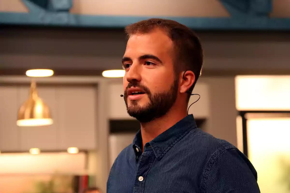 Un hombre con barba y micrófono de diadema, vestido con una camisa azul, está hablando en un entorno interior iluminado con luces cálidas.