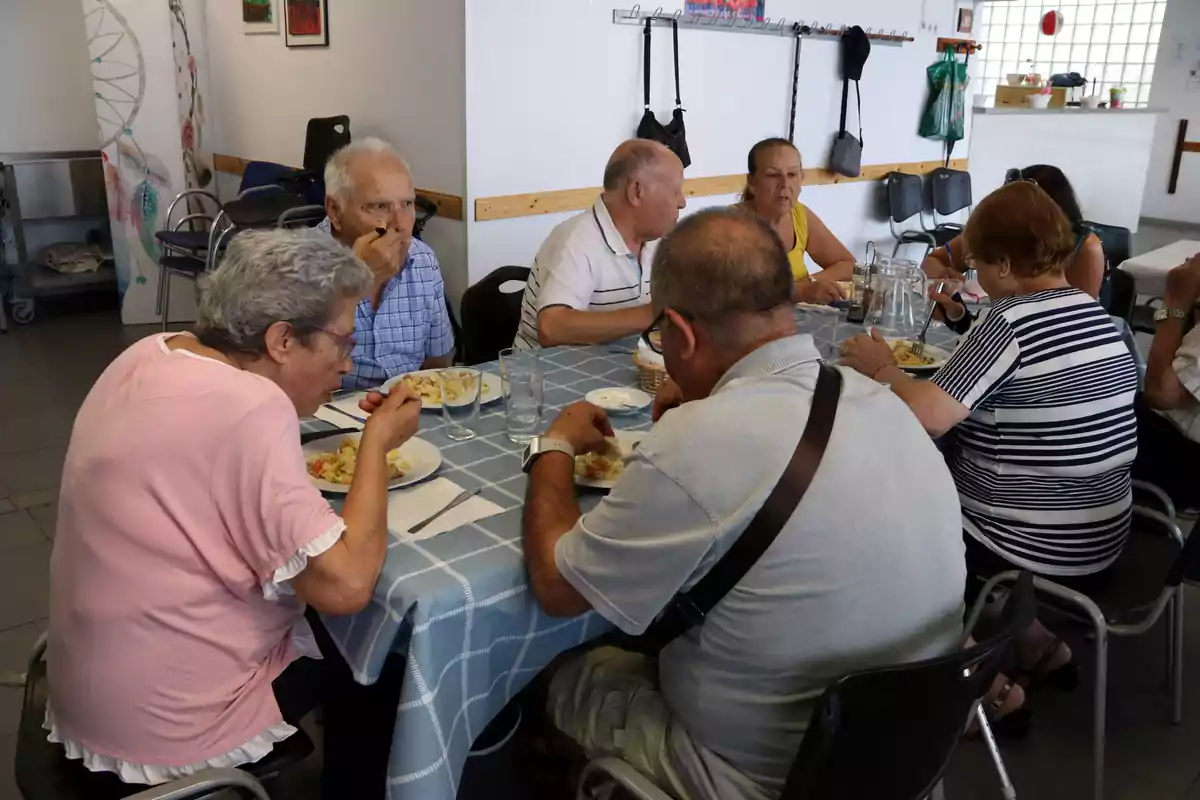 Un grup de persones grans compartint un dinar en una taula amb estovalles blaves en un menjador comunitari.