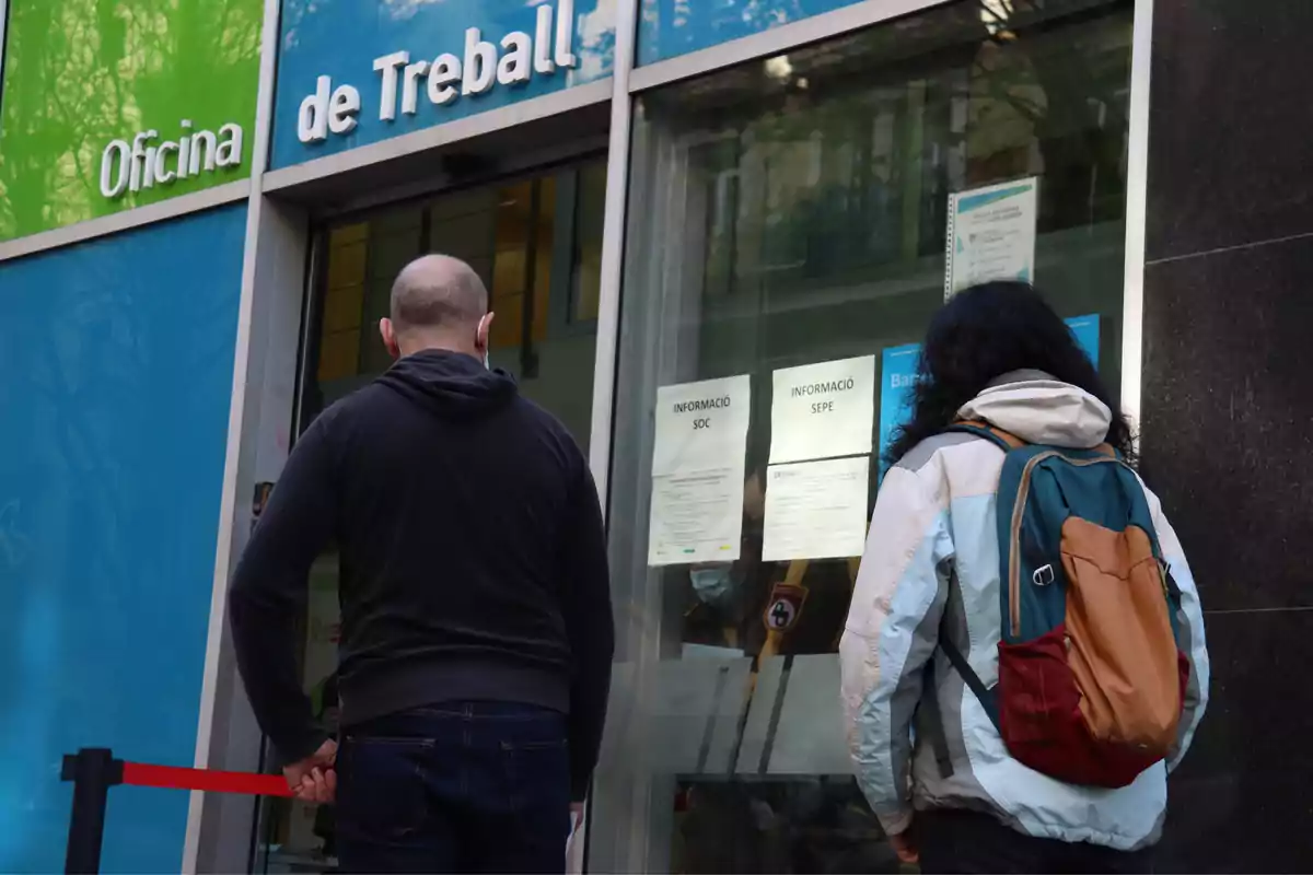 Dos personas están de pie frente a la entrada de una oficina de empleo llamada "Oficina de Treball" en un edificio con fachada azul y verde.